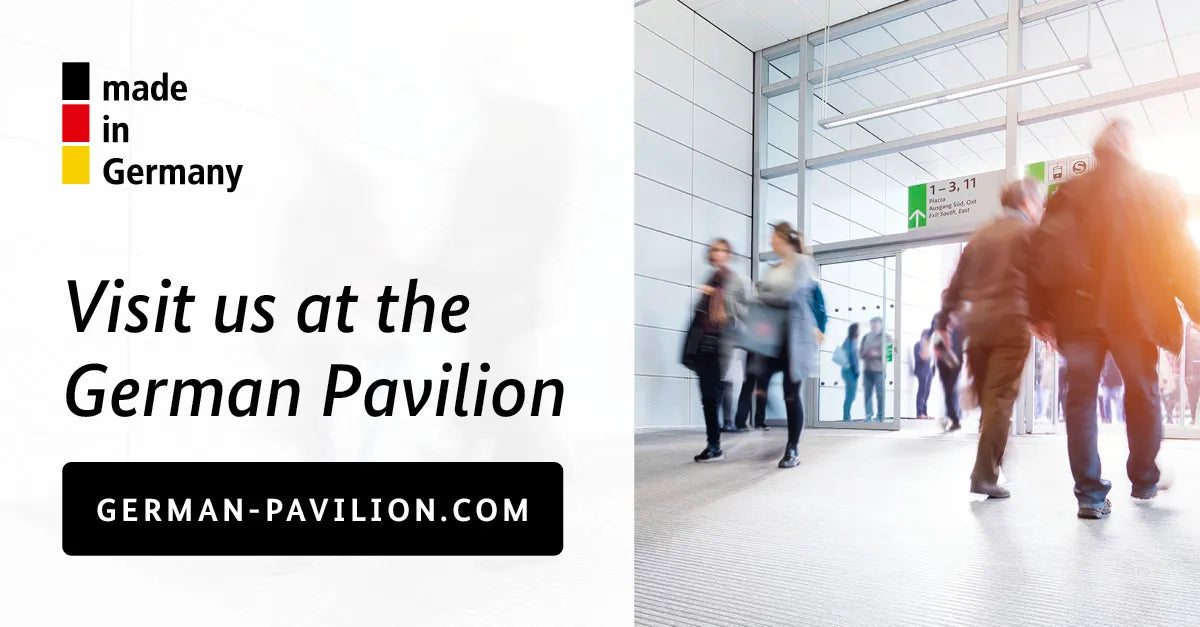 Eingangsbereich einer Messehalle mit mehreren vorbeigehenden Besuchern; links steht der Schriftzug „made in Germany“ in Schwarz-Rot-Gold sowie „Visit us at the German Pavilion“ und darunter die Webadresse german-pavilion.com.
