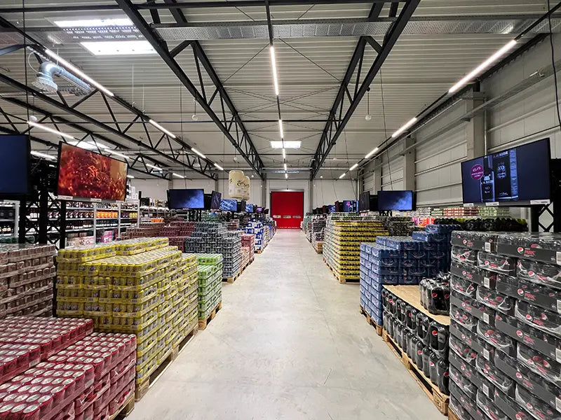Innenaufnahme eines großflächigen Getränkemarktes mit hohen Regalen und palettenweise gestapelten Dosen und Flaschen; breite Mittelgänge, helle LED-Beleuchtung an der Decke sowie mehrere Bildschirme mit Werbegrafiken entlang der Verkaufsfläche.