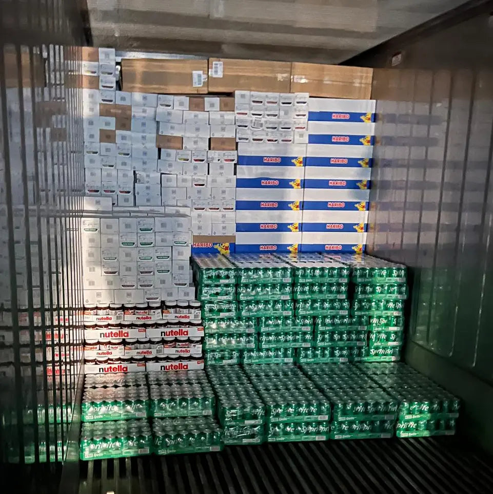 Innenansicht eines Lkw-Aufliegers, beladen mit palettenweise gestapelter Ware; unten mehrere grüne Getränkedosen, darüber Kartons mit Nutella, Haribo und weiteren Markenprodukten, sauber und dicht aneinandergereiht für den Transport.