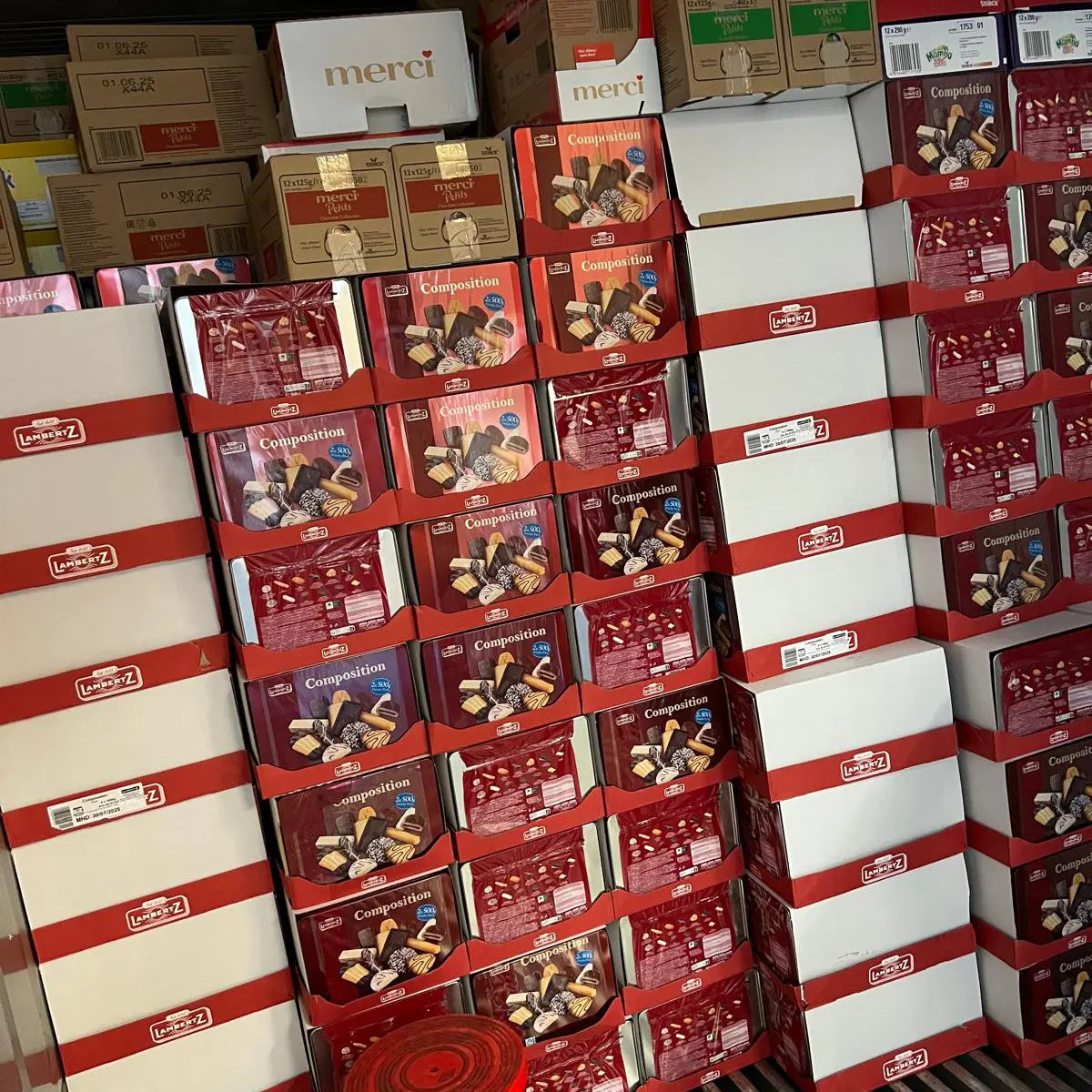 Innenansicht eines Lkw-Aufliegers, dicht bepackt mit gestapelten Süßwarenkartons; vorne zahlreiche rote Lambertz-„Composition“-Displays, darüber Kartons von Merci sowie weitere gemischte Waren, sauber und stabil bis zur Decke geschichtet.
