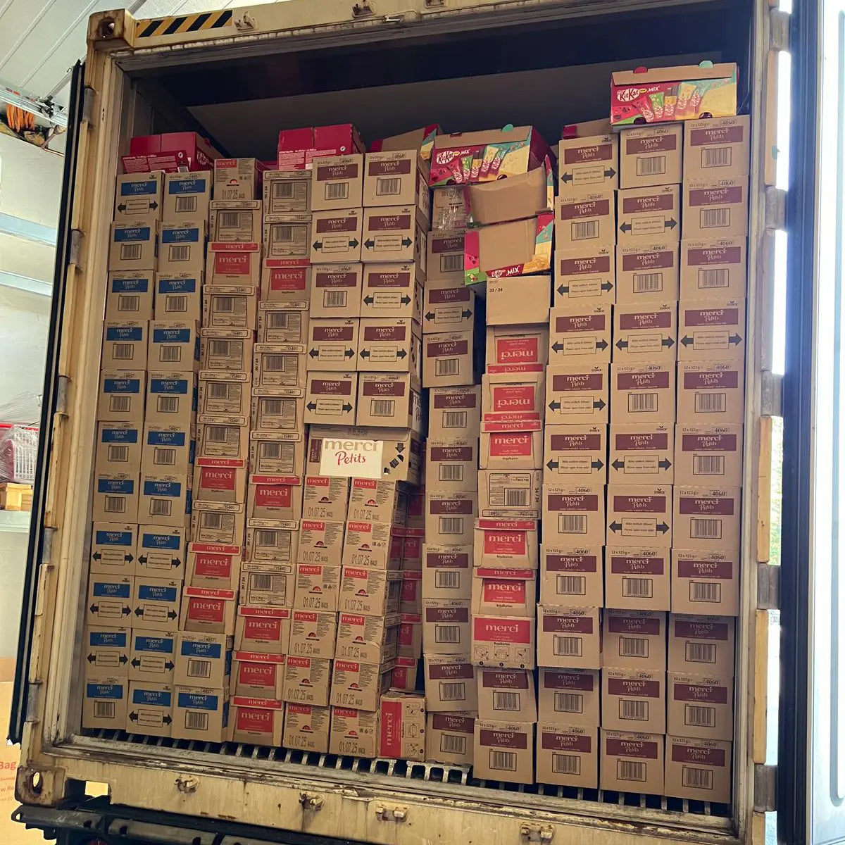 Innenansicht eines voll beladenen Lkw-Aufliegers mit dicht gestapelten Kartons verschiedener Süßwarenmarken, darunter zahlreiche Merci-Kartons und bunte Mischkartons von KitKat; die Ware ist bis unter die Decke gestapelt.